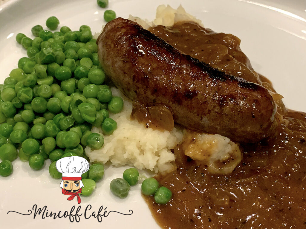 One sausage link laying on a bed of mashed potatoes, smothered with brown onion gravy and a pile of green peas on a white plate. 