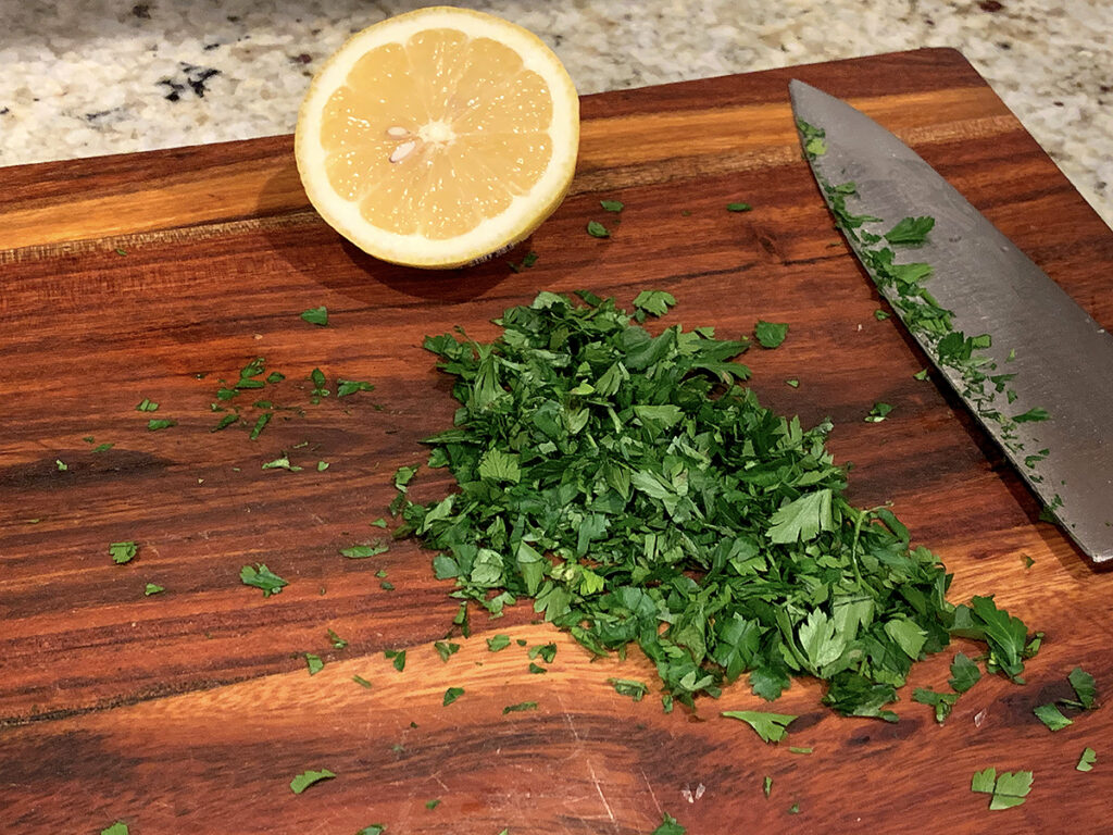 One half of a lemon, a pile of chopped parsley, and a chef knife on a wood cutting board. 
