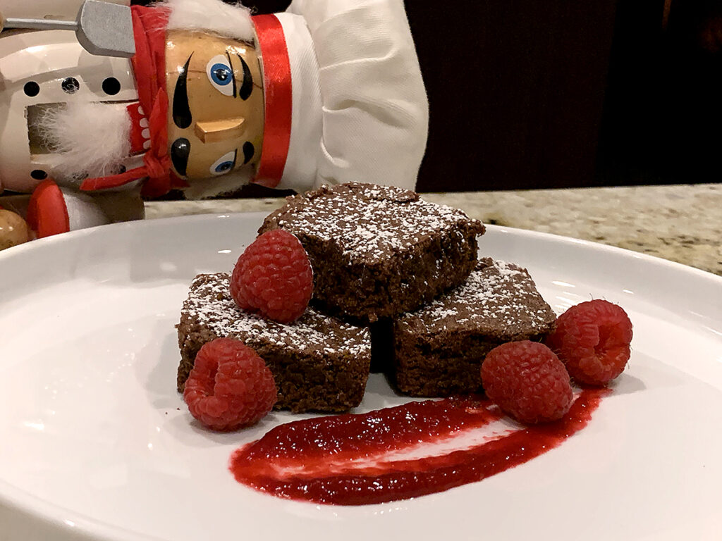 Three fudge brownies stacked on a white plate, dusted with powdered sugar and garnished with a swoosh of red raspberry sauce as well as fresh raspberries. There's also a nutcracker who looks like chef in the background.