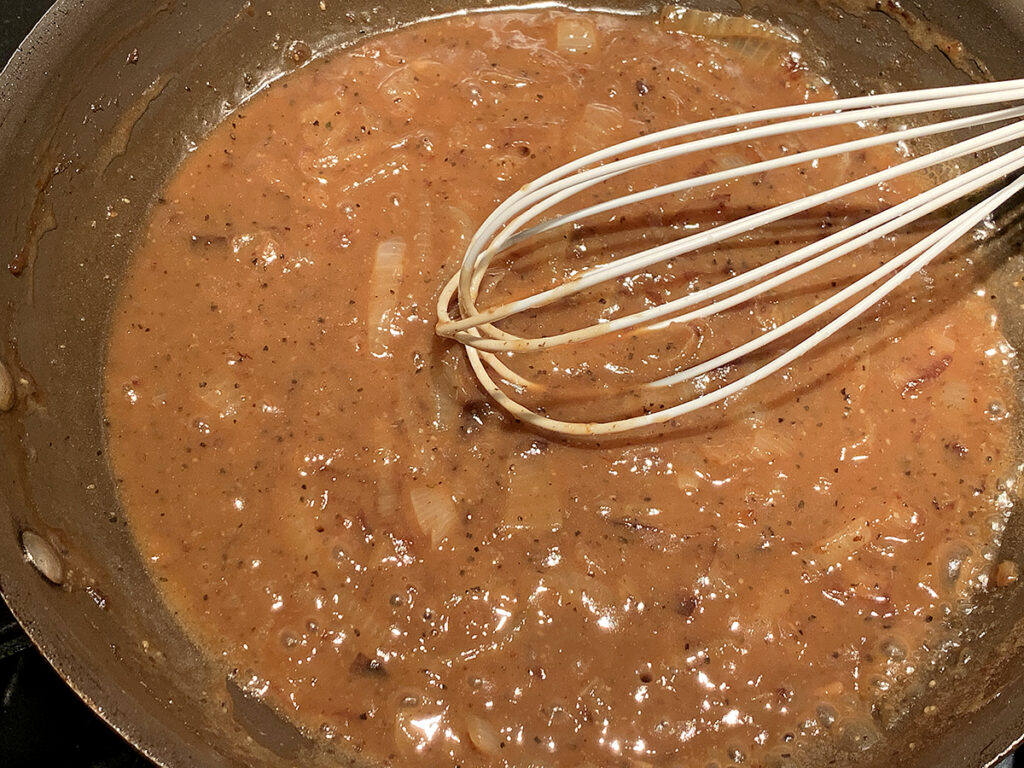 Dark brown gravy and onions simmering in a skillet and being stirred with a white rubber whisk. 