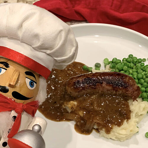 One sausage link laying on a bed of mashed potatoes, smothered with brown onion gravy and a pile of green peas on a white plate. There's a nutcracker in the foreground who looks like a chef.