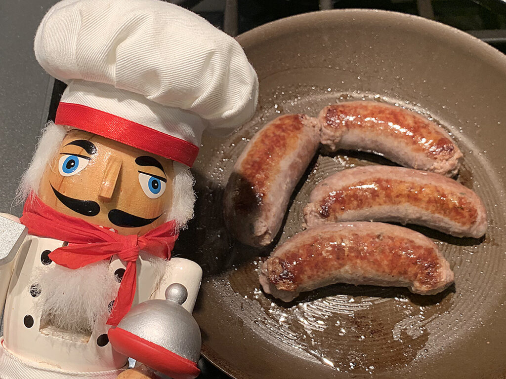 Four plum sausages browning in a skillet. There's a nutcracker in the foreground who looks like a chef.