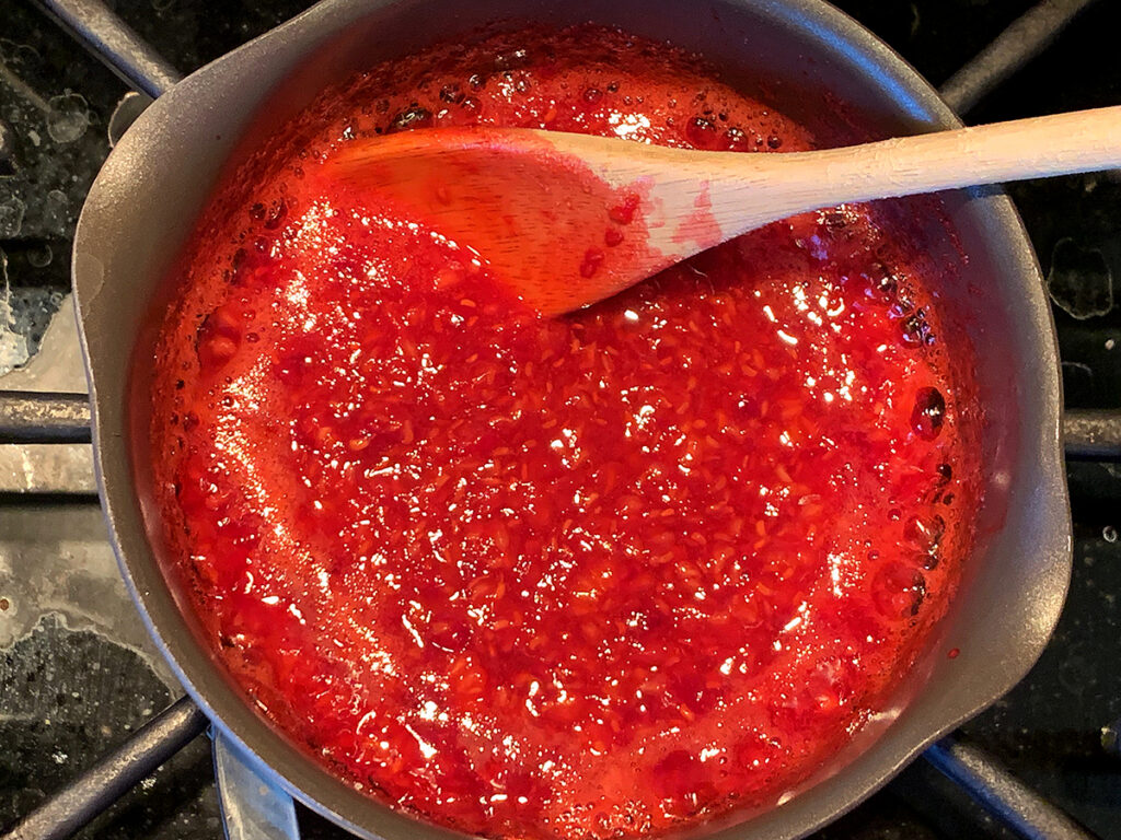 Overhead view of broken down raspberries simmering in a saucepan with a wooden spoon.