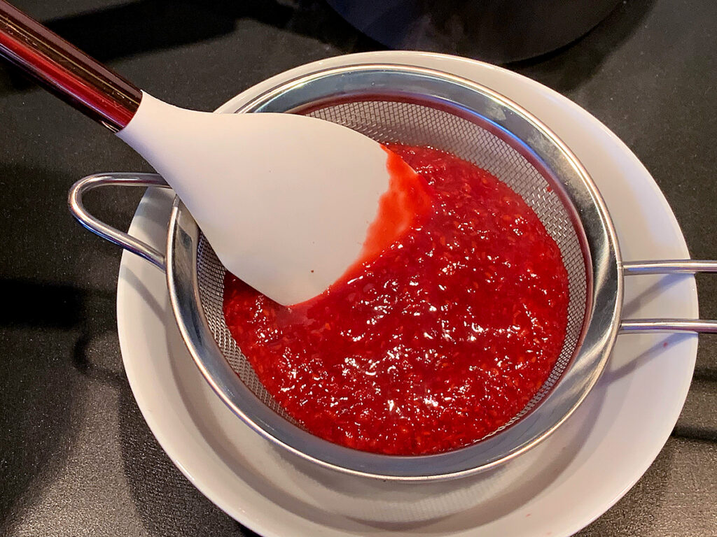 Straining raspberry sauce in a sieve to remove seeds with a rubber spatula.