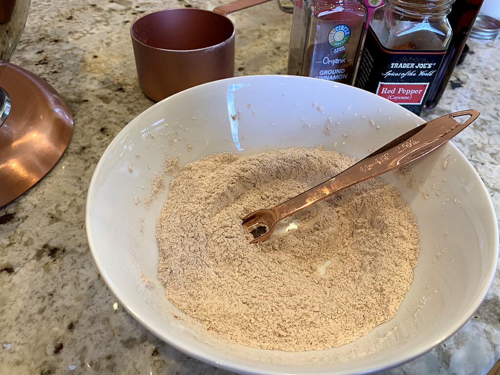 Tan mixture of flour, cayenne pepper and cinnamon in a small white bowl being stirred with a copper measuring spoon.