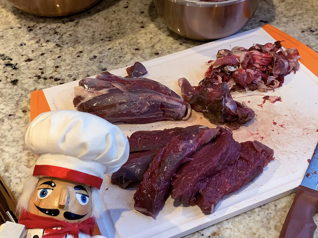 Several large pieces of dark red deer meat on a white cutting board, removing silver skin. There's a nutcracker in the foreground who looks like a chef.