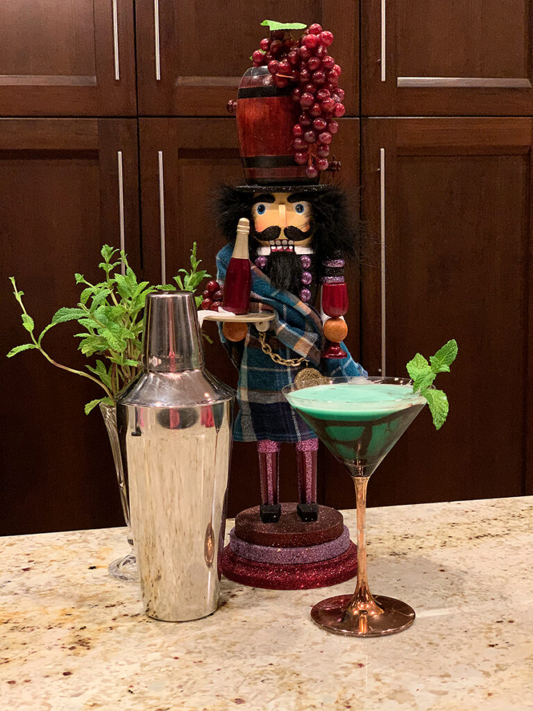 Green cocktail in a copper stemmed martini glass and garnished with mint leaves and chocolate drizzle. There's also silver a cocktail shaker, ice bucket and a nutcracker who looks like a sommelier.