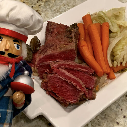 A platter of corned beef, cabbage and carrots with a nutcracker who looks like a chef and is wearing a kilt in the foreground.