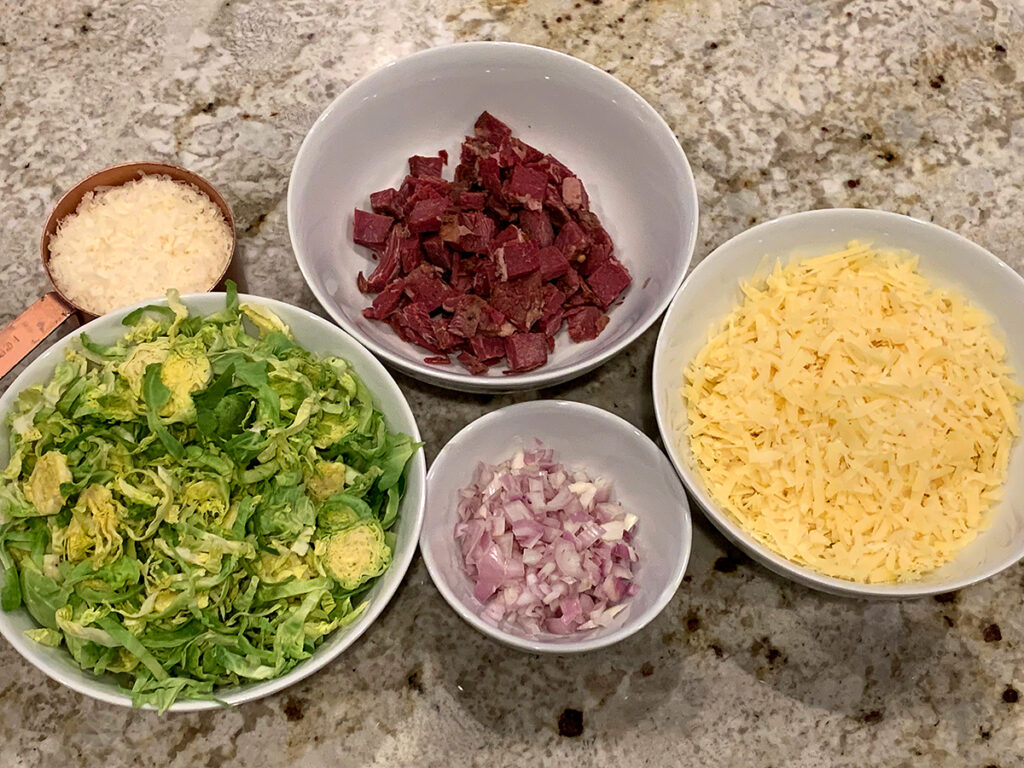 Shredded brussels sprouts, cubed corned beef, shredded white cheese, minced shallots and parmesan cheese all in separate bowls. 