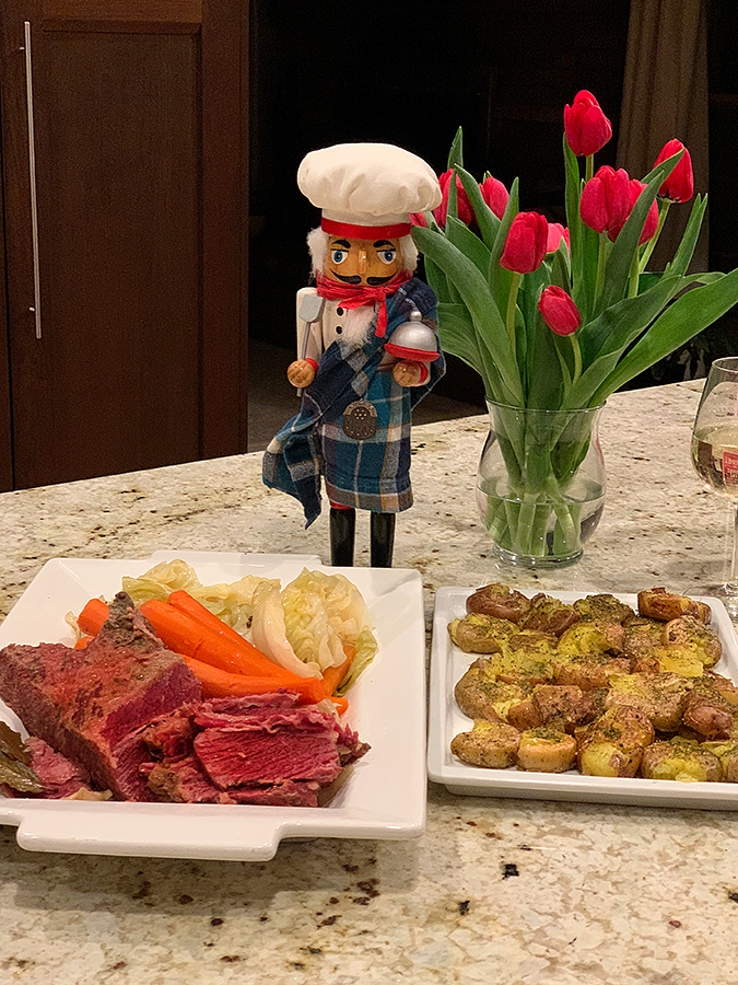 A platter with corned beef, cabbage and carrots. Another platter has small baby potatoes that are just flattened slightly and sprinkled with dill weed. There's a nutcracker who looks like a chef and wearing a kilt, in the background, next to a vase of red tulips.