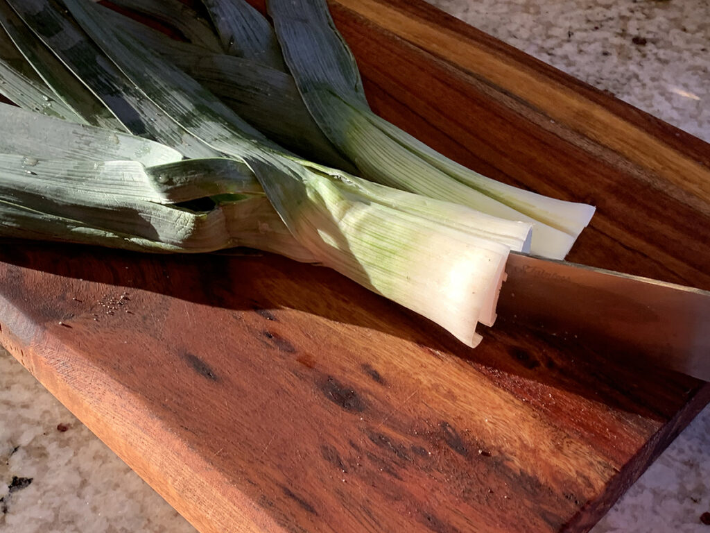 Two leeks on a wood cutting board. The root ends are cut off and they are being cut vertically. 