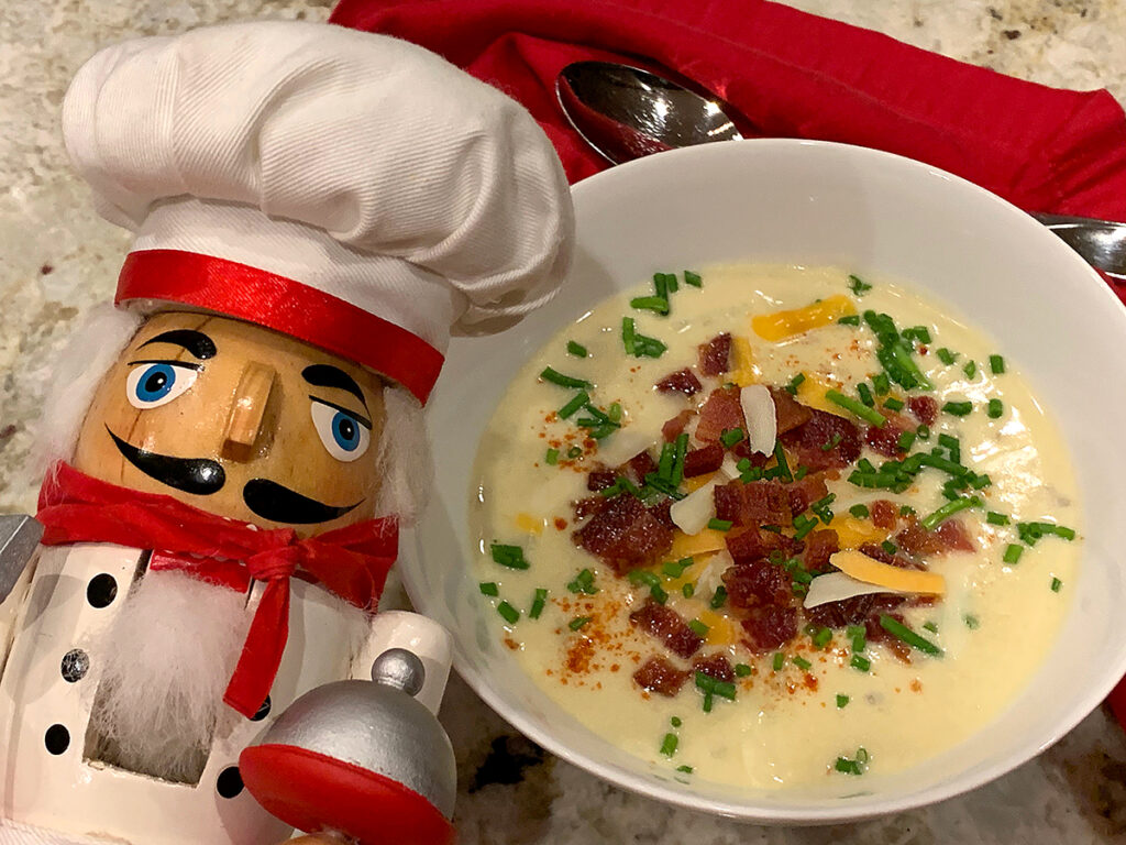 Pale yellow soup in a round white bowl, topped with shredded cheese, chopped bacon and chives. There's a nutcracker in the foreground who looks like a chef.