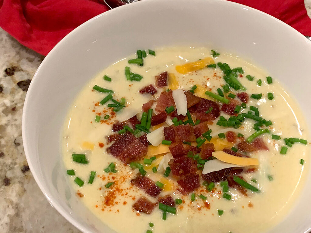 Pale yellow soup in a round white bowl, topped with shredded cheese, chopped bacon and chives.