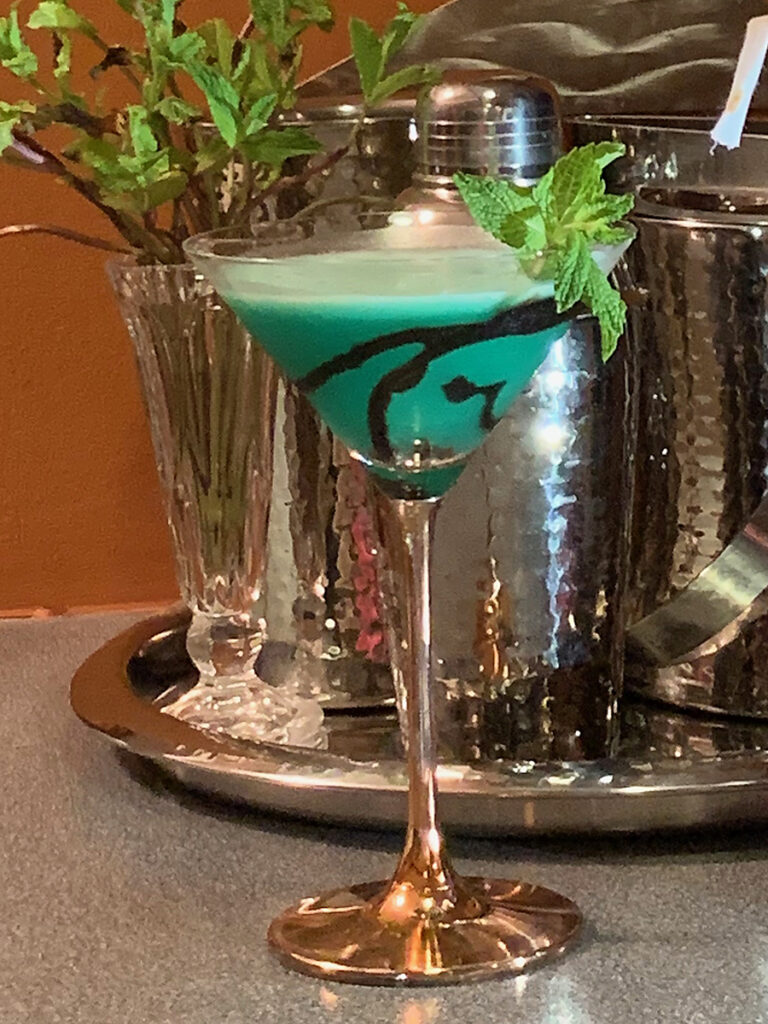 Green cocktail in a copper stemmed martini glass and garnished with mint leaves and chocolate drizzle. There's also silver a cocktail shaker, ice bucket.