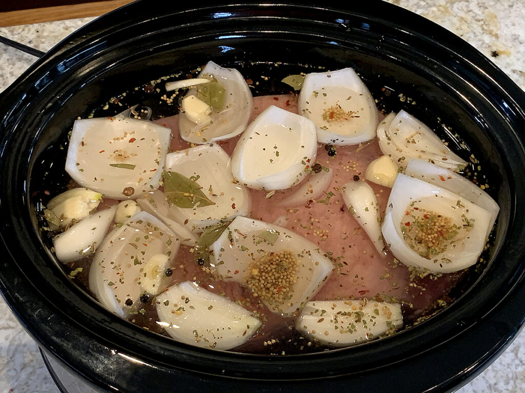 Corned beef in a slow cooker, covered with water and with large chunks of onions, some bay leaves, garlic cloves and peppercorns on top.