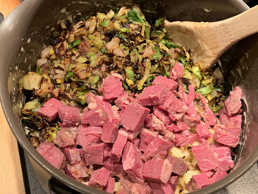 Cooked shredded brussels sprouts and cubed cooked corned beef being added to the saucepan with the mac and cheese. 