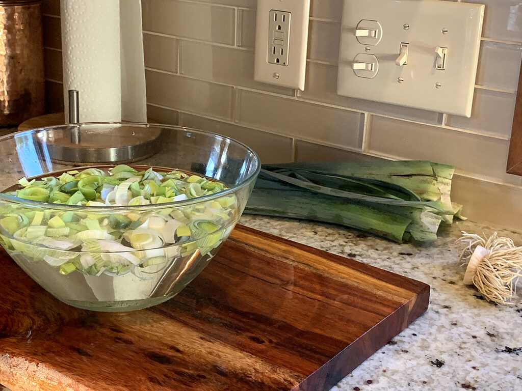 Chopped leeks rinsed in a clear glass bowl sitting on a wood board. The dark part of the leeks are laying in the background. 
