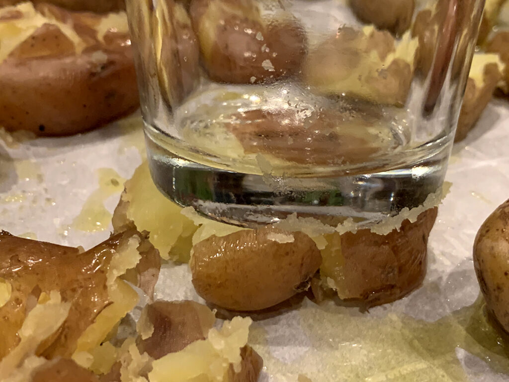 Close up of bottom of pint glass slightly smashing baby potatoes on a sheet pan. 