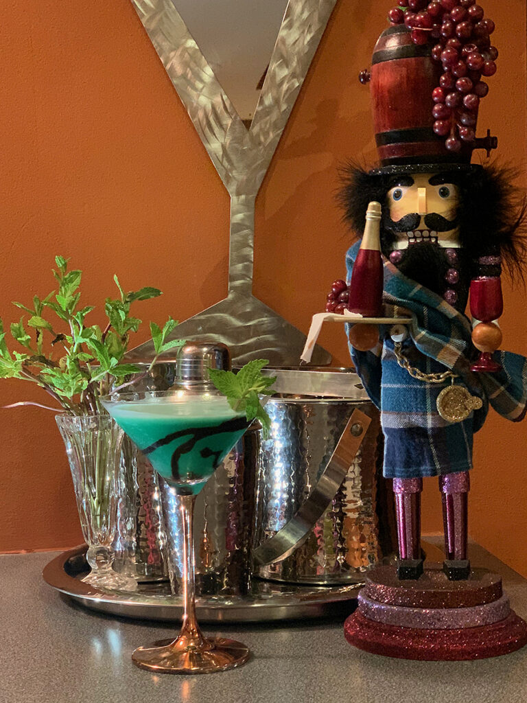 Green cocktail in a copper stemmed martini glass and garnished with mint leaves and chocolate drizzle. There's also silver a cocktail shaker, ice bucket and a nutcracker who looks like a sommelier. 