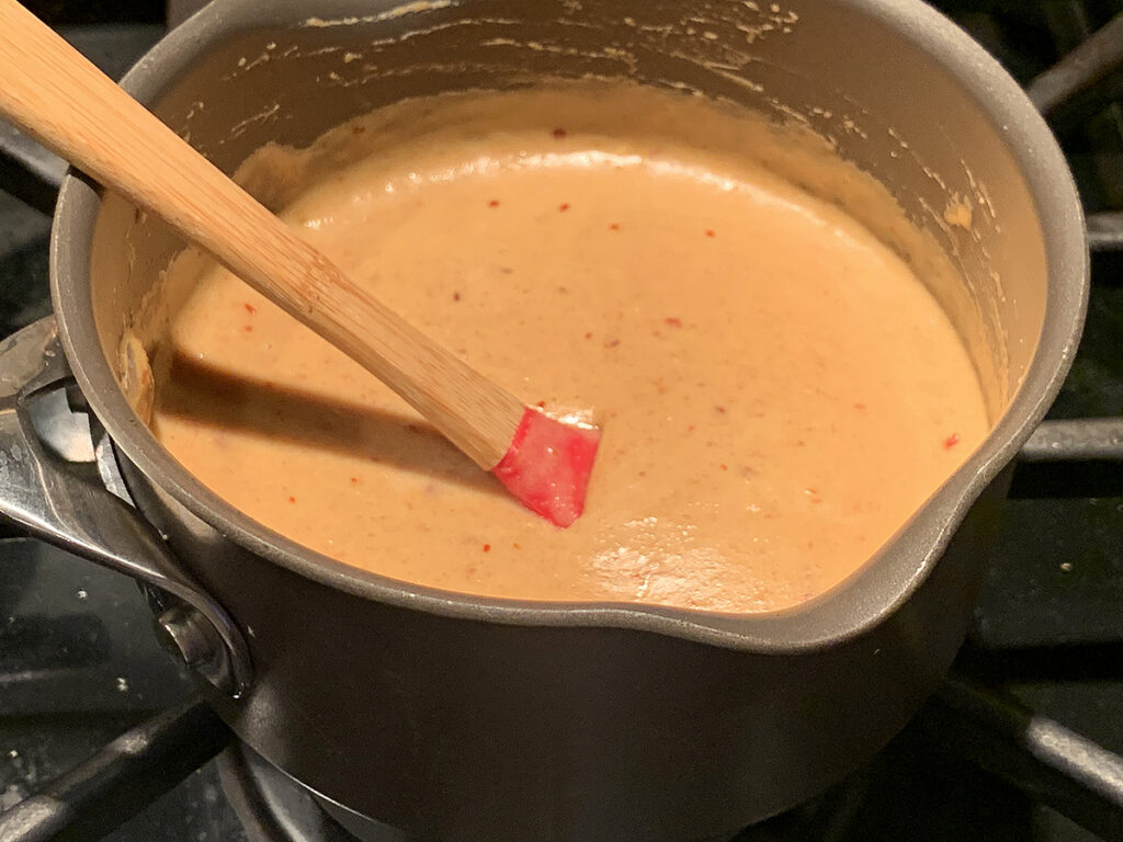creamy tan sauce in a small sauce pan being stirred with a wooden spatula with a red rubber tip.