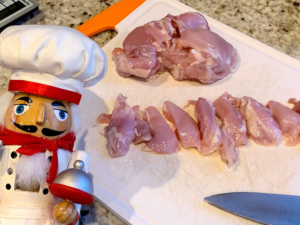 Boneless, skinless chicken thighs being cut into strip on a white plastic cutting board. There's a nutcracker who looks like a chef in the foreground.