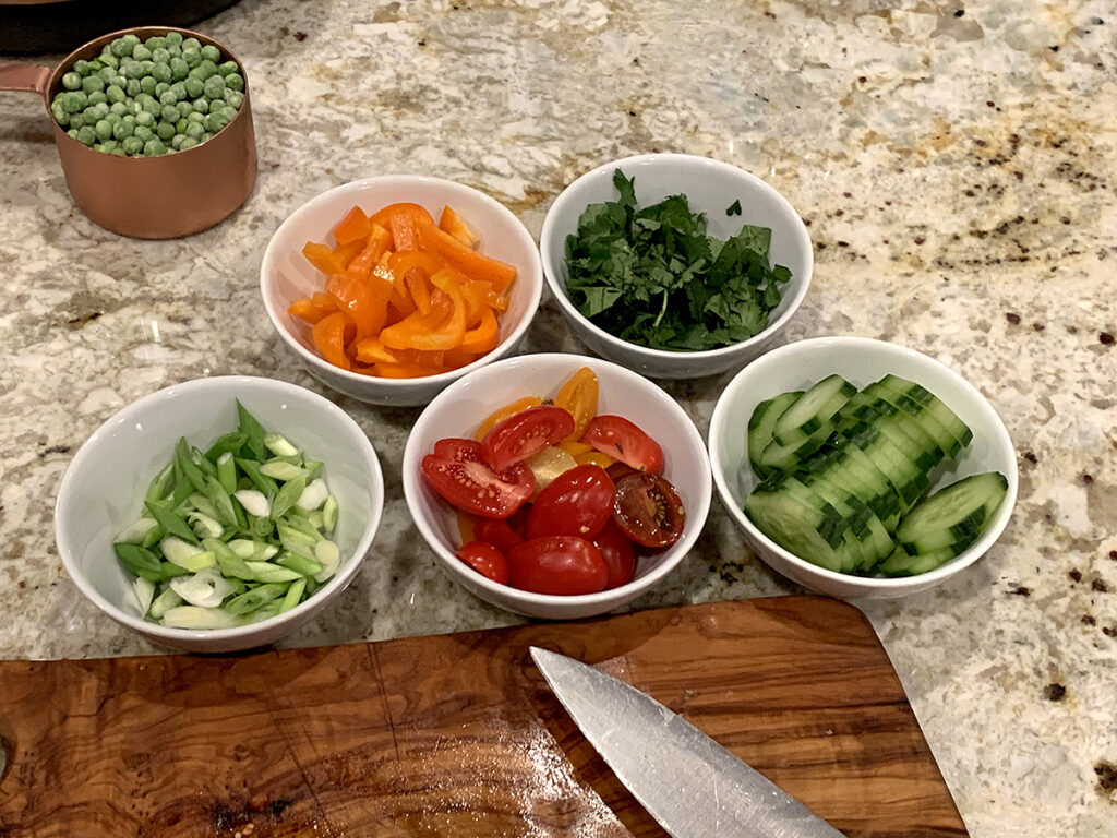 Five small round white bowls with separate ingredients (orange bell peppers, cilantro, chopped green onions, red cherry tomato haves and sliced cucumbers) on a granite counter top. There's also a copper measuring cup filled with frozen green peas.