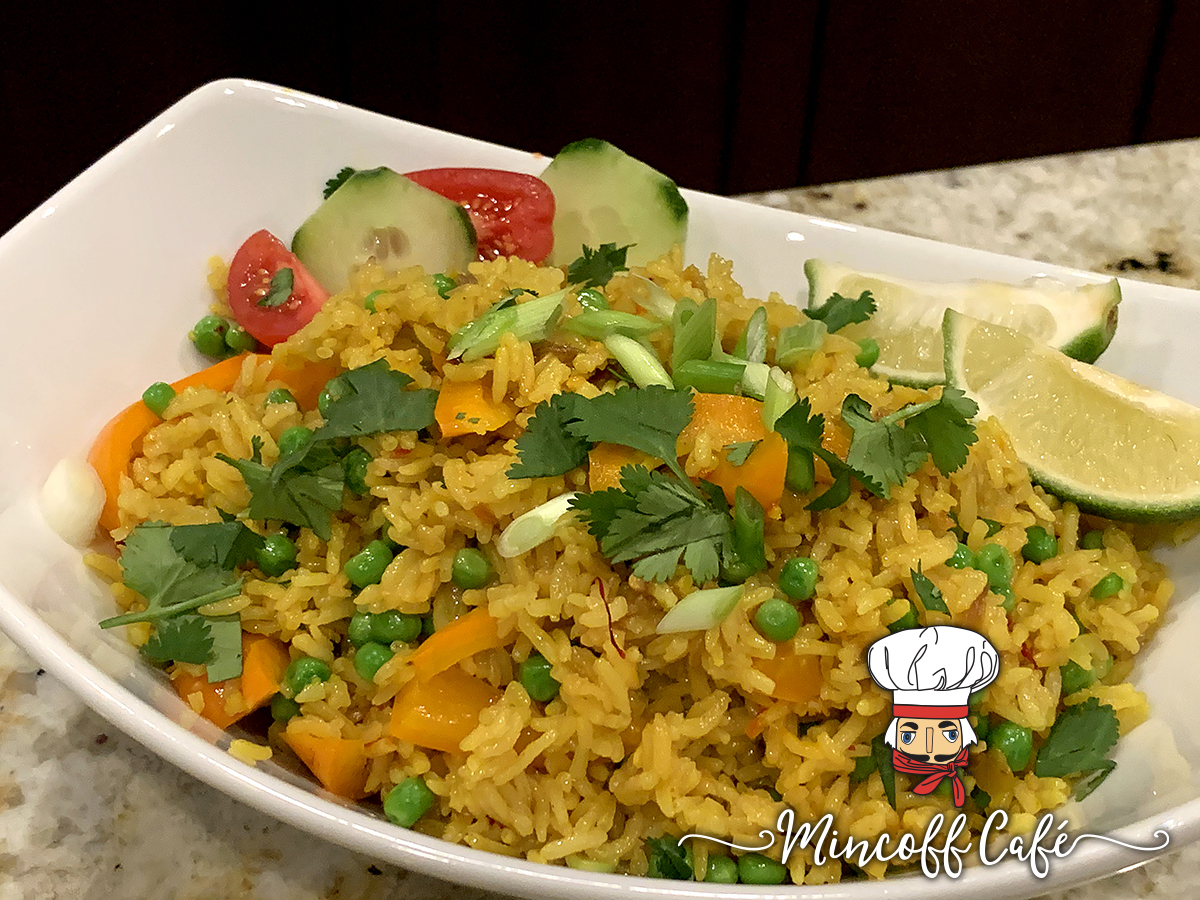 White oval bowl with yellow rice, garnished with lime wedges, cucumber slices, tomato slices, orange bell peppers, chopped green onions and cilantro.