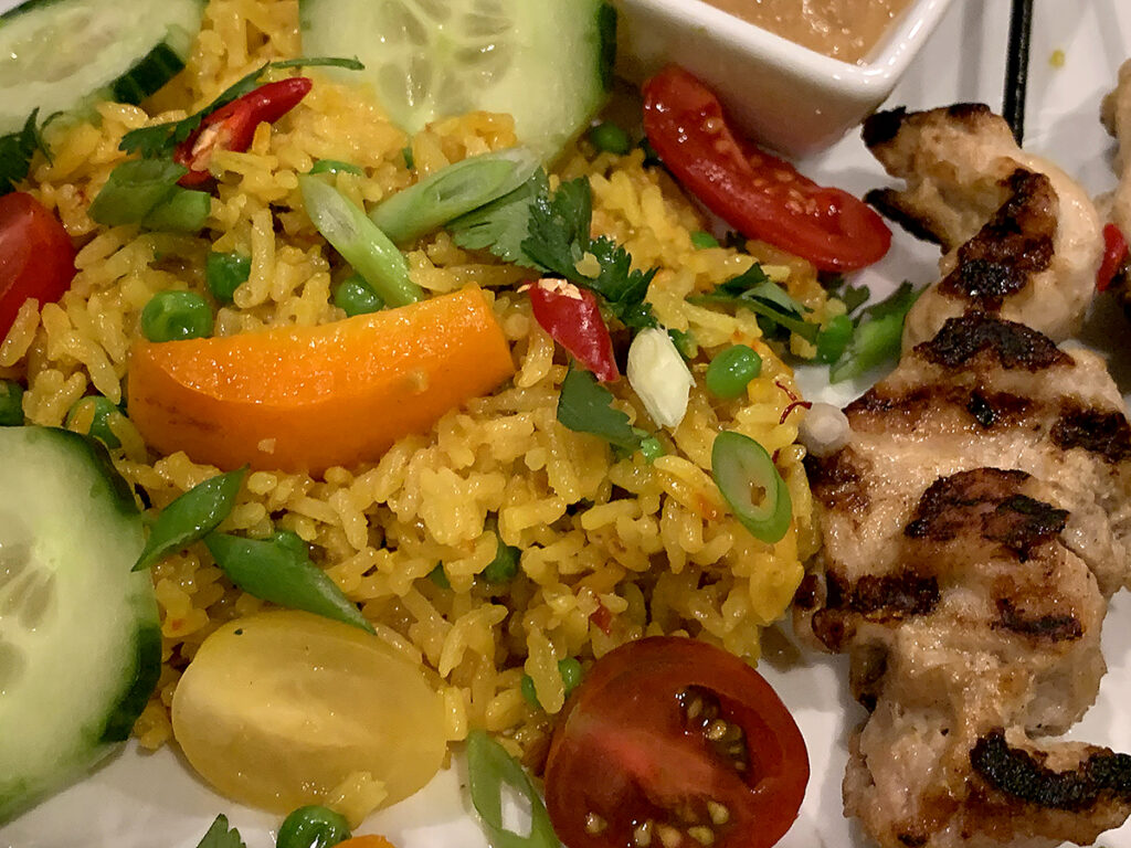 Close up of two skewers of charred, grilled chicken, yellow rice with cucumber, tomato and orange bell pepper slices.