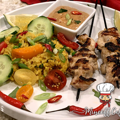 Two skewers of charred, grilled chicken, yellow rice with cucumber, tomato and orange bell pepper slices, on a round white plate along with a tan sauce in a square white ramekin and all garnished with lime wedges, red Thai chilies and cilantro.