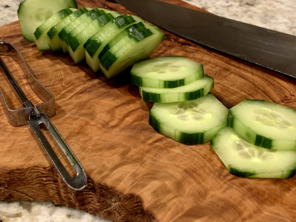 Cucumber slices on a wood cutting board, along with a vegetable peeler and a chef knife.