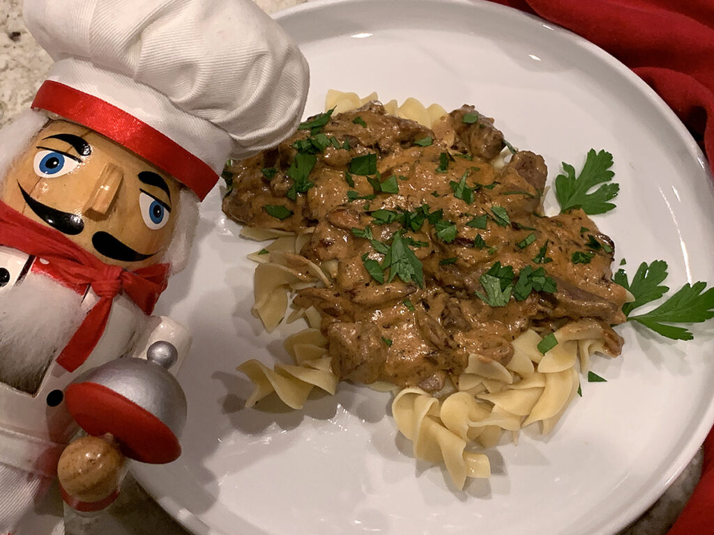 Light brown slightly chunky sauce on top of pale tan wide egg noodles on a white plate. It's sprinkled with green bits of parsley and there's a nutcracker in the foreground that looks like a chef.