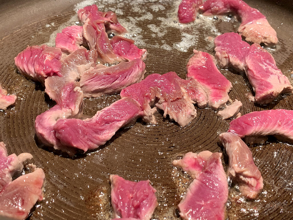 Thin strips of rare venison searing in a skillet.