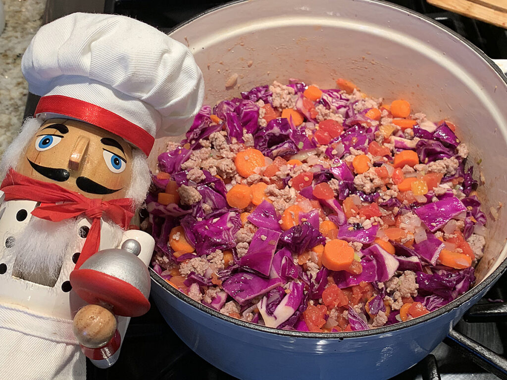 Bright purple cabbage and orange carrots mixed with browned ground beef in a white dutch oven. There's a nutcracker in the foreground who looks like a chef.