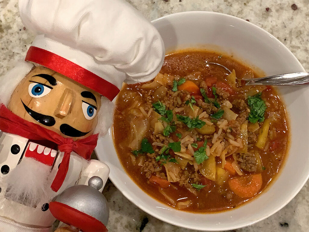 A bowl of cabbage roll soup with it's red broth and garnished with green parsley. There's a nutcracker in the foreground who looks like a chef.