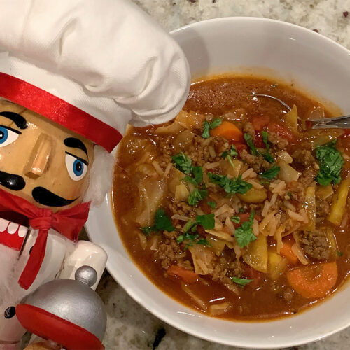 A bowl of cabbage roll soup with it's red broth and garnished with green parsley. There's a nutcracker in the foreground who looks like a chef.
