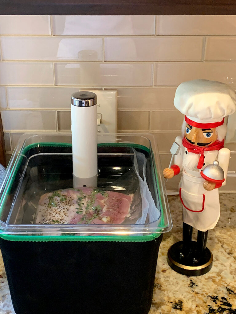 Large plastic and insulated square container filled with water, vacuum sealed bags of turkey, a sous vide device, with a nutcracker who looks like a chef standing by.