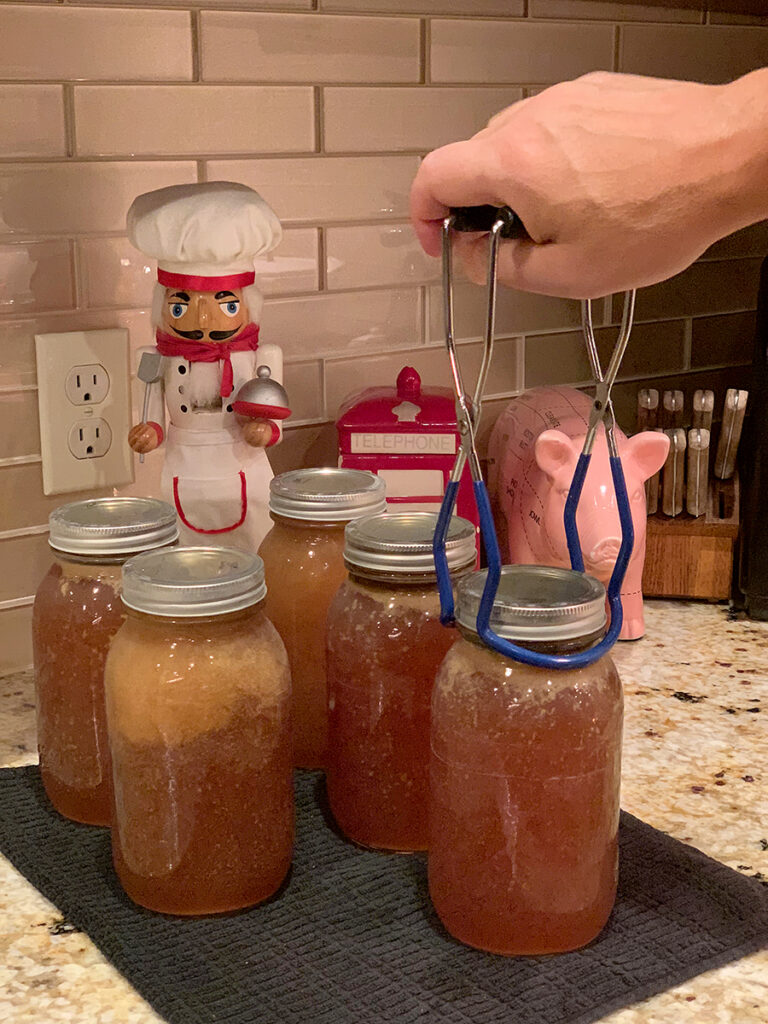 Several large glass jars filled with a dark amber liquid. One is being placed via canning tongs. There's a nutcracker in the background who looks like a chef.