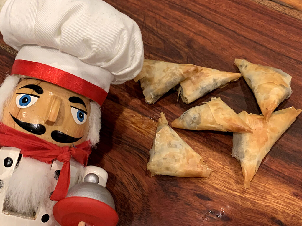 Five lightly browned triangle phyllo dough packets sitting on a wood cutting board. There's a nutcracker in the foreground who looks like a chef.