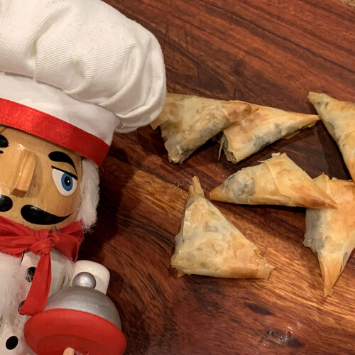 Five lightly browned triangle phyllo dough packets sitting on a wood cutting board. There's a nutcracker in the foreground who looks like a chef.