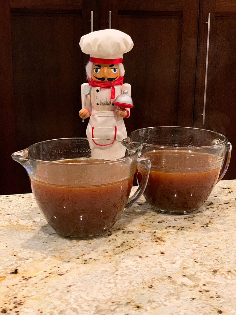 Two very large glass measuring cups fill with dark brown turkey stock. There's a nutcracker in the background who looks like a chef.