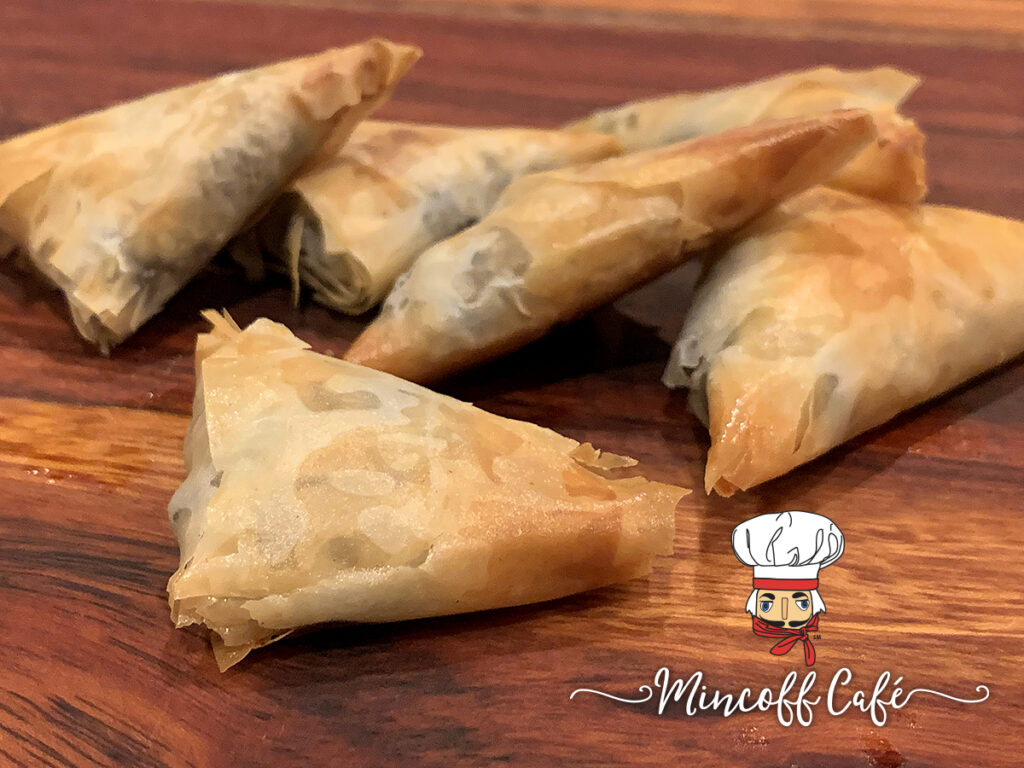Five lightly browned triangle phyllo dough packets sitting on a wood cutting board.