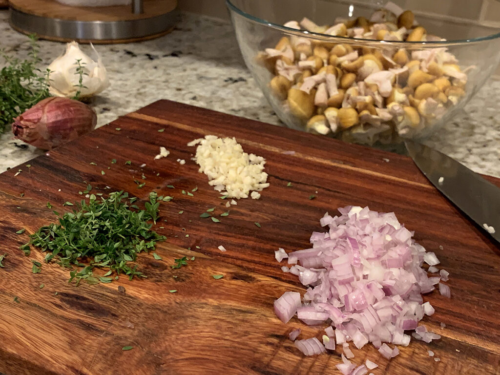 Chopped shallots, garlic and thyme on a wood cutting board. There's a clear bowl of chopped yellow mushrooms in the background.
