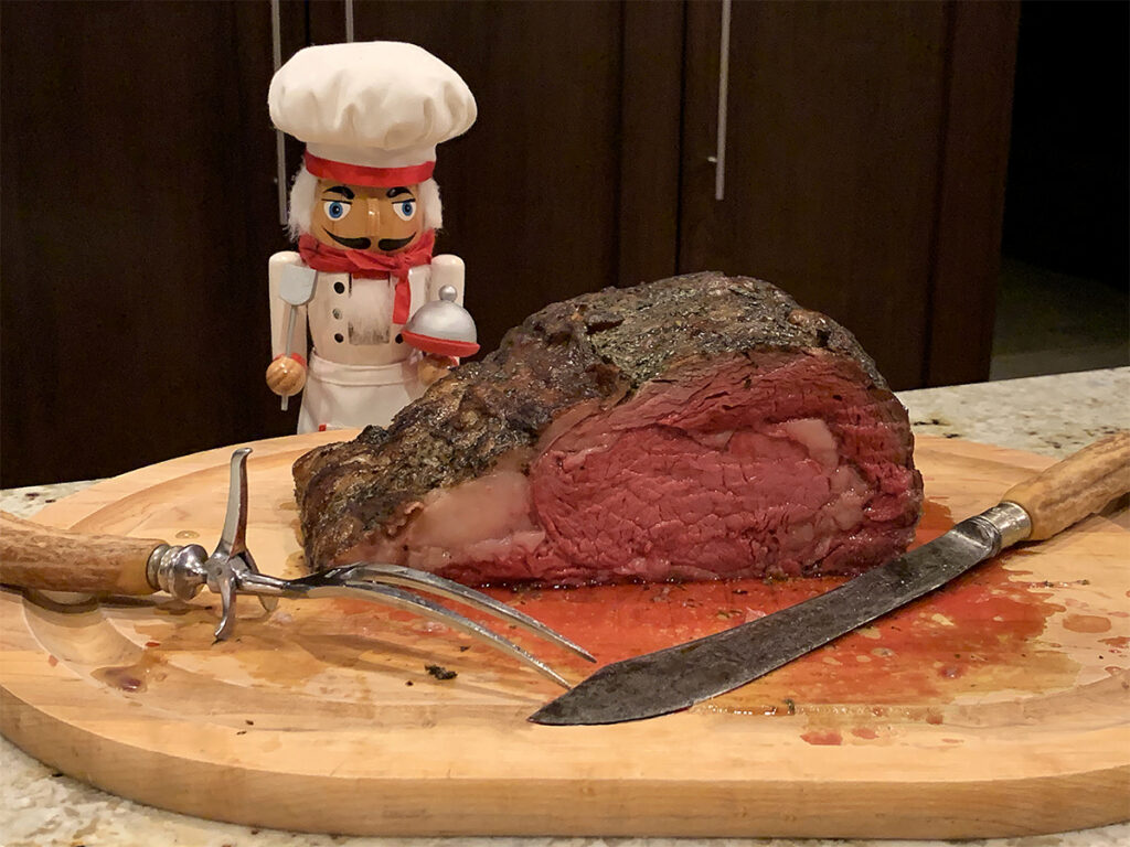 Juicy redish pink prime rib roast on wood cutting board with vintage knife and fork, and a nutcracker who looks like a chef in the background.