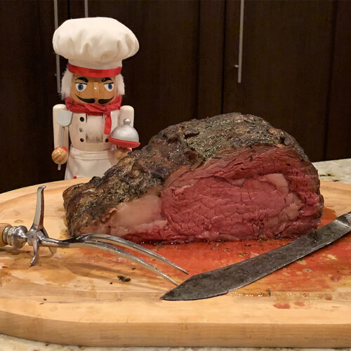 Juicy redish pink prime rib roast on wood cutting board with vintage knife and fork, and a nutcracker who looks like a chef in the background.