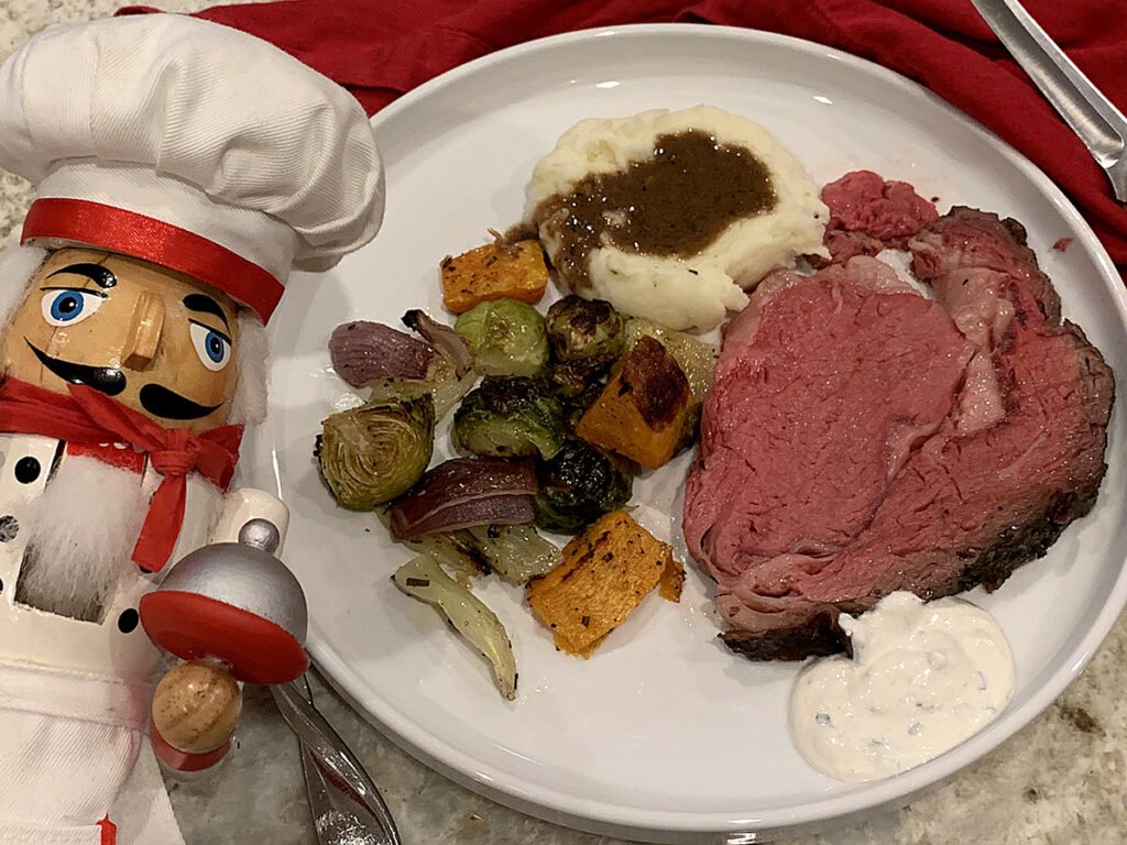 Round white dinner plate with rare rib roast, mashed potatoes & gravy, and roasted veggies. There's a nutcracker in the foreground who looks like a chef.