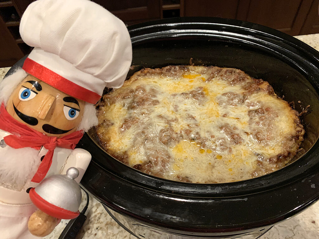 Lasagna cooked in a Crockpot. There's a nutcracker in the foreground who looks like a chef. 