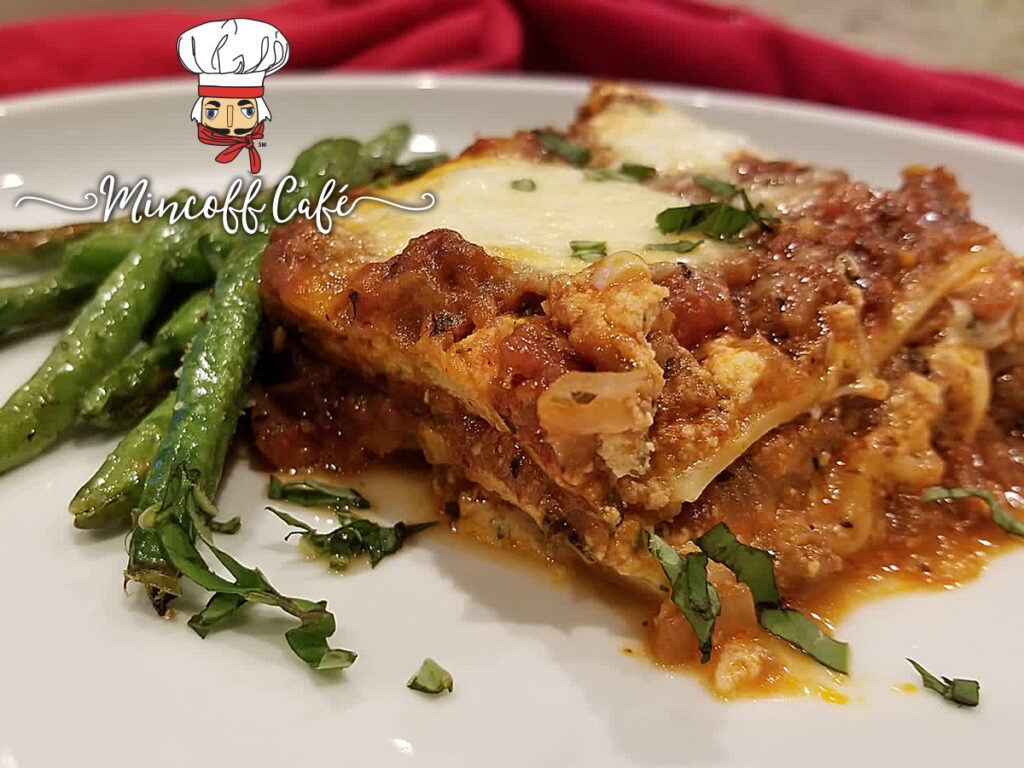 Slice of cheesy lasagna with red meat sauce on a white plate with green beans.