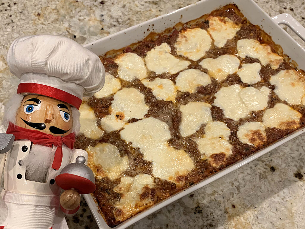 Baked cheesy lasagna in a white rectangular casserole dish . There's a nutcracker in the foreground who looks like a chef.
