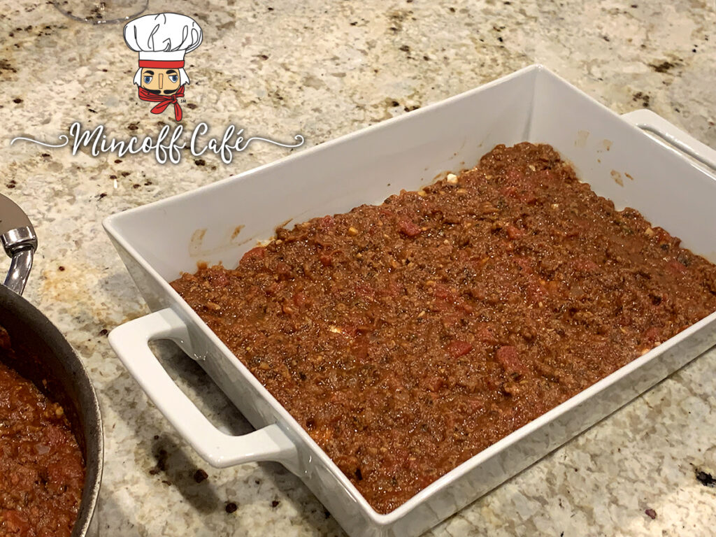A ayer of the red meat sauce is on the bottom of a white rectangular casserole dish.