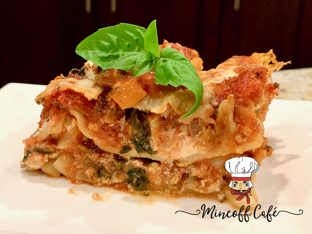 Slice of cheesy vegetarian lasagna with red meat sauce on a white plate garnished with a bright green tip of fresh basil.